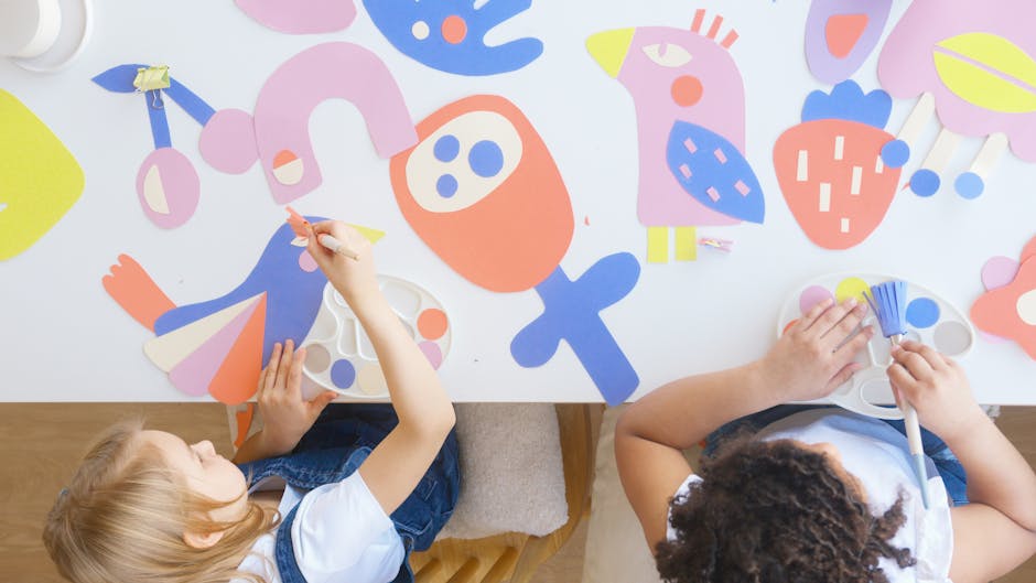 Two children at a table working on colorful paper art cutouts with paintbrushes.